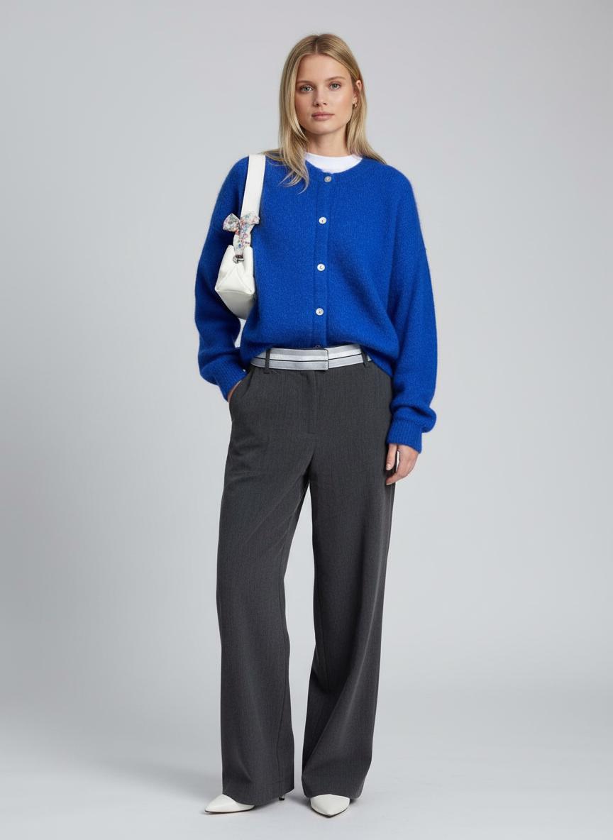 Woman wearing a blue cardigan and gray pants with a white handbag on a gray background