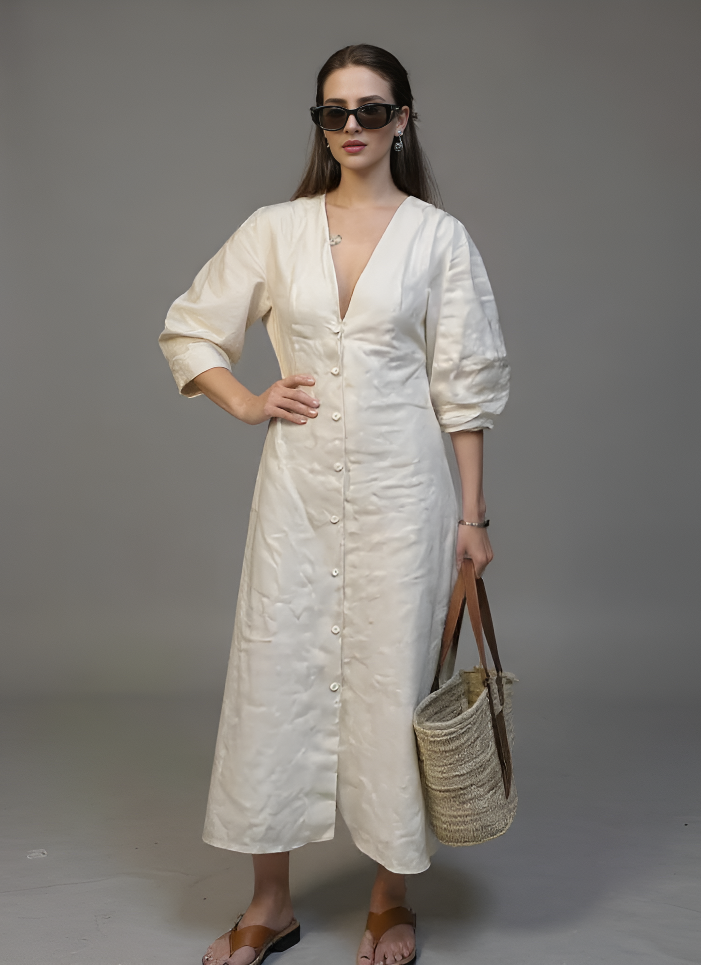 Woman in a white dress holding a straw bag against a gray background