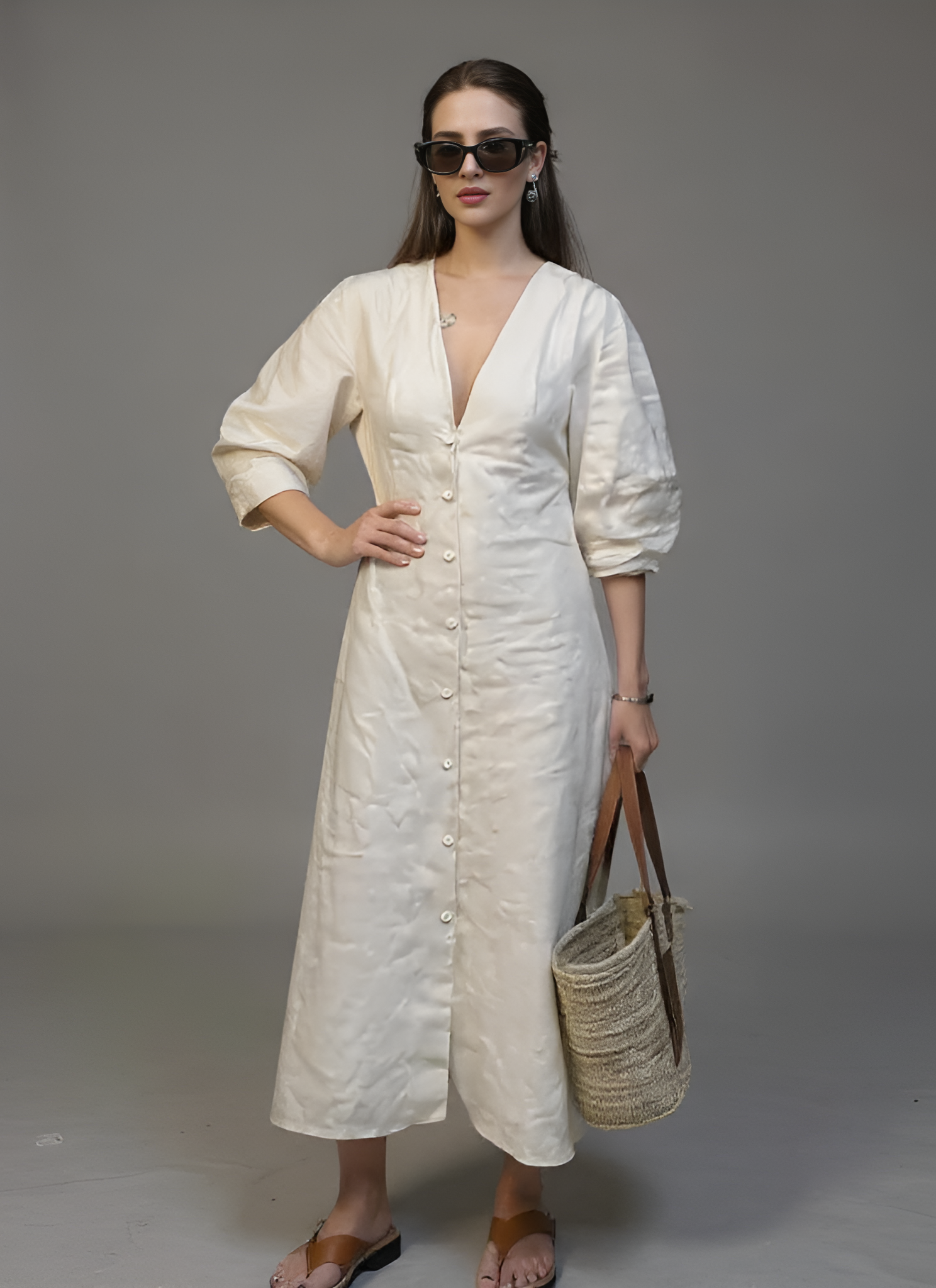 Woman in a white dress holding a straw bag against a gray background
