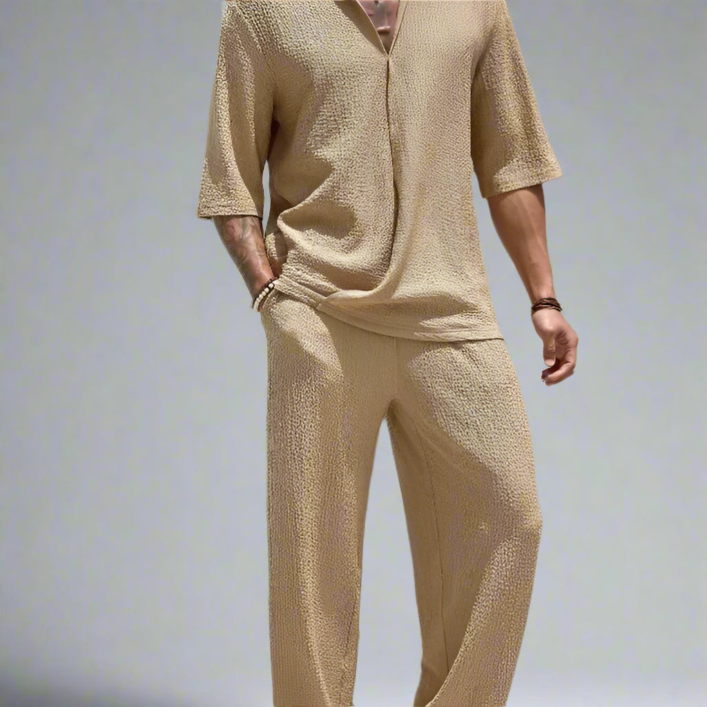 Person wearing a beige textured outfit standing outdoors with a white wall and greenery in the background.