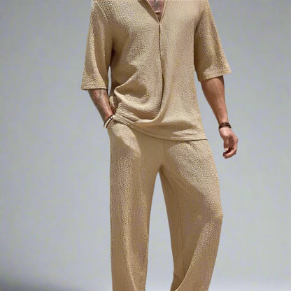 Person wearing a beige textured outfit standing outdoors with a white wall and greenery in the background.